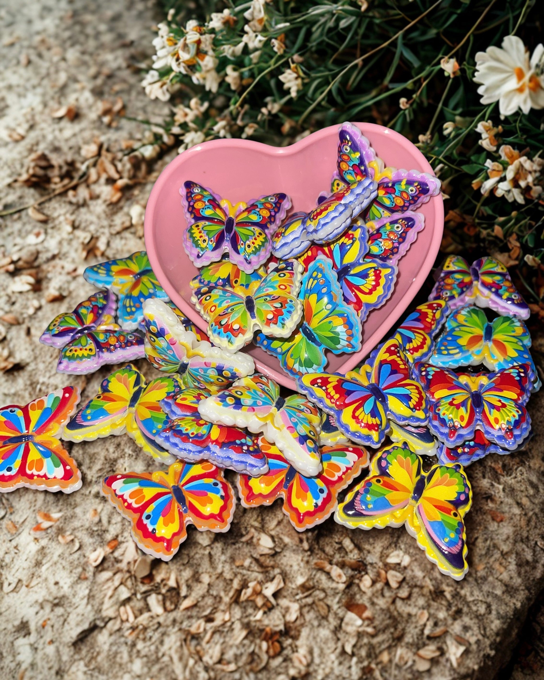 Colorful butterfly beads in a pink heart bowl on stone surface with wildflowers